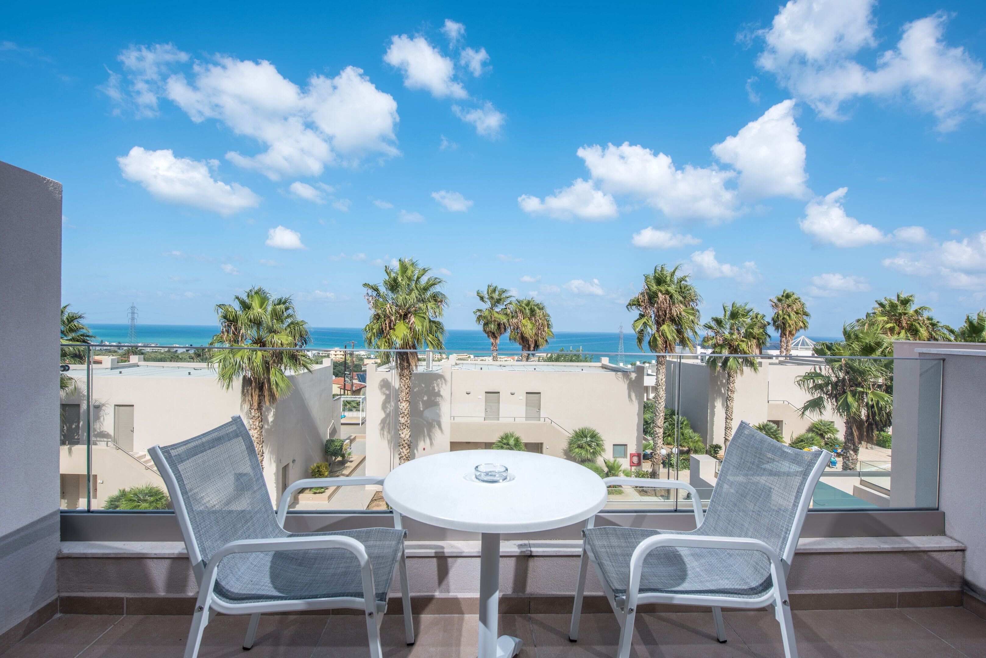 Un balcón con mesa y dos sillas blancas, con vista a una playa y palmeras.