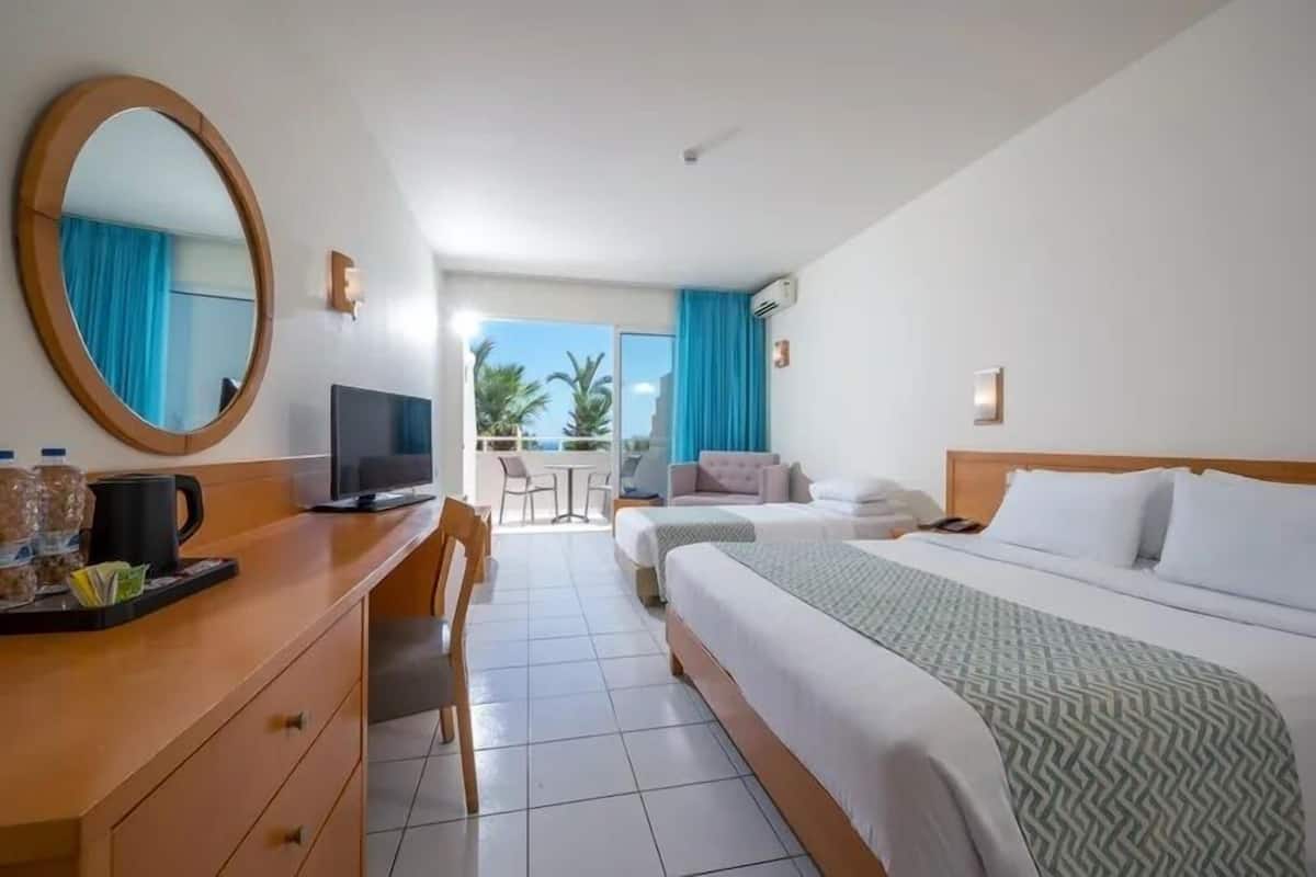 Habitación de hotel con cama, escritorio con computadora, silla, televisor y ventana con vista a palmeras.
