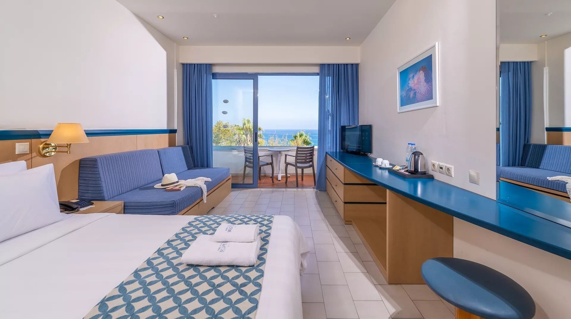 A hotel room with a bed, a sofa, a desk, a chair, a TV, and a balcony with a view.
