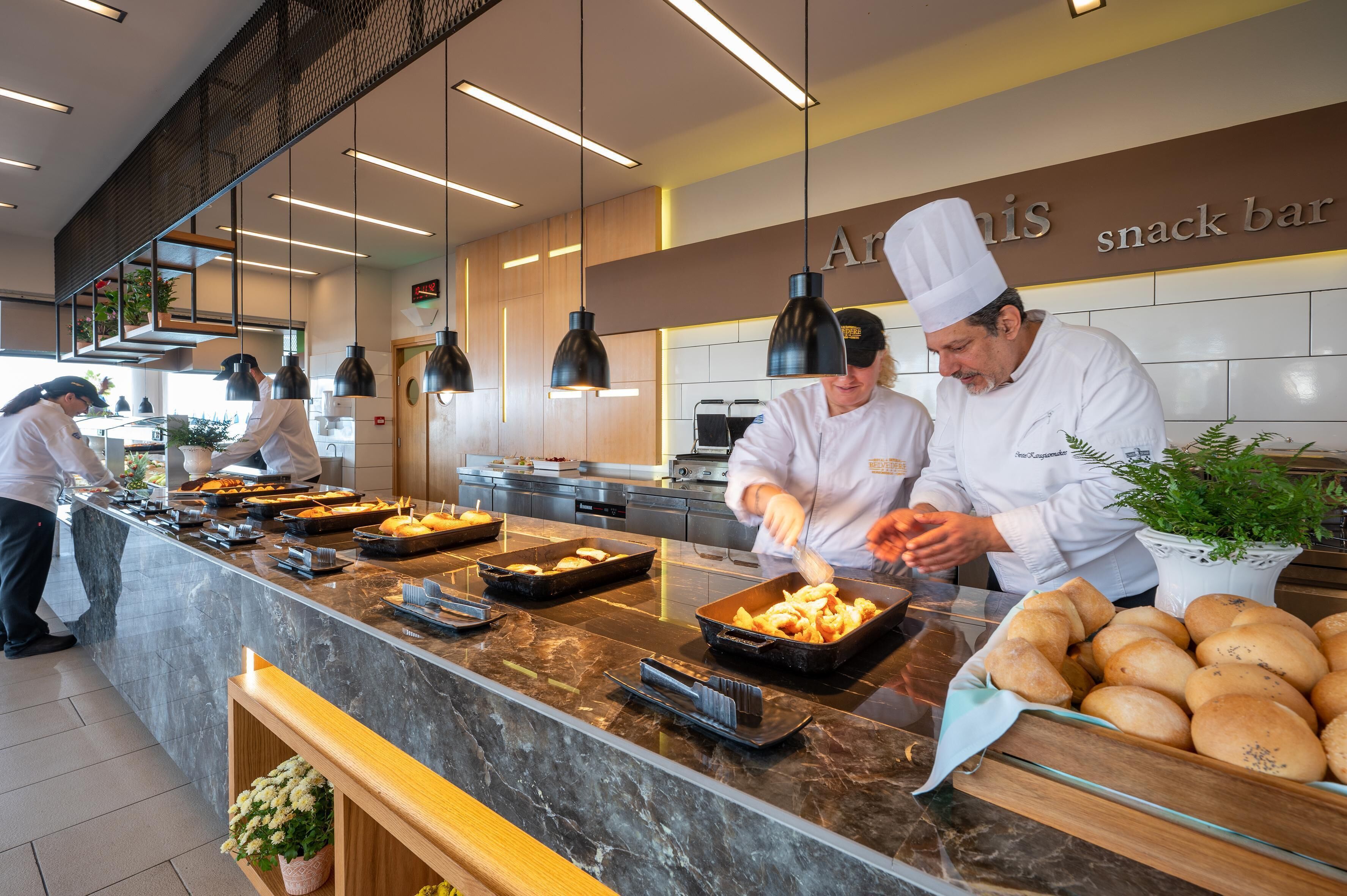 A modern kitchen with chefs preparing food, a marble countertop, and a sign for 'Aris snack bar'.