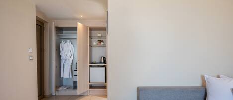A modern hotel room with a grey sofa, a white pillow, a robe hanging in the closet, and a small kitchenette.