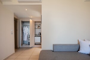 Quarto de hotel moderno com sofá cinzento, almofada branca, roupão pendurado no armário e uma pequena cozinha equipada.