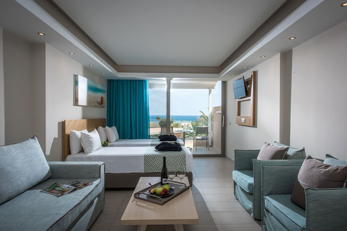 Habitación de hotel con una cama, dos sofás, una mesa de centro con fruta y copas de vino, y un televisor en la pared.
