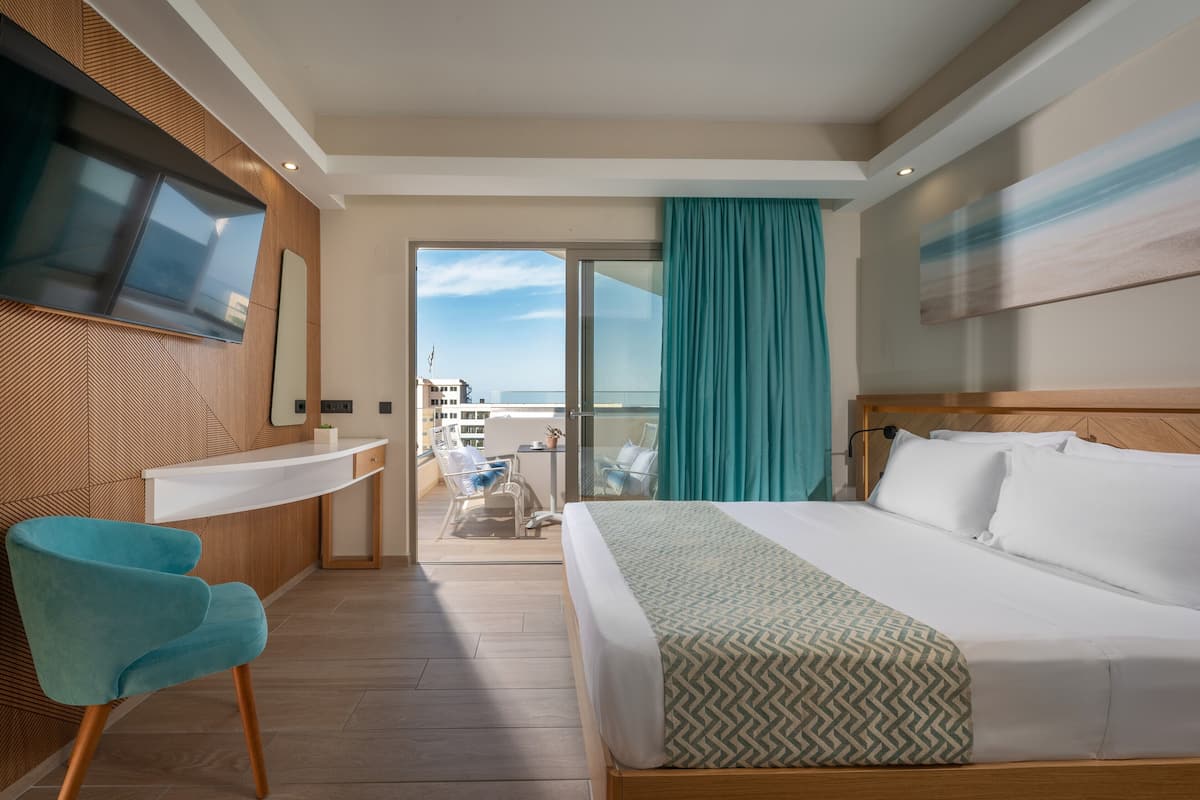 Habitación de hotel con cama, escritorio, silla, televisor y balcón con vistas.