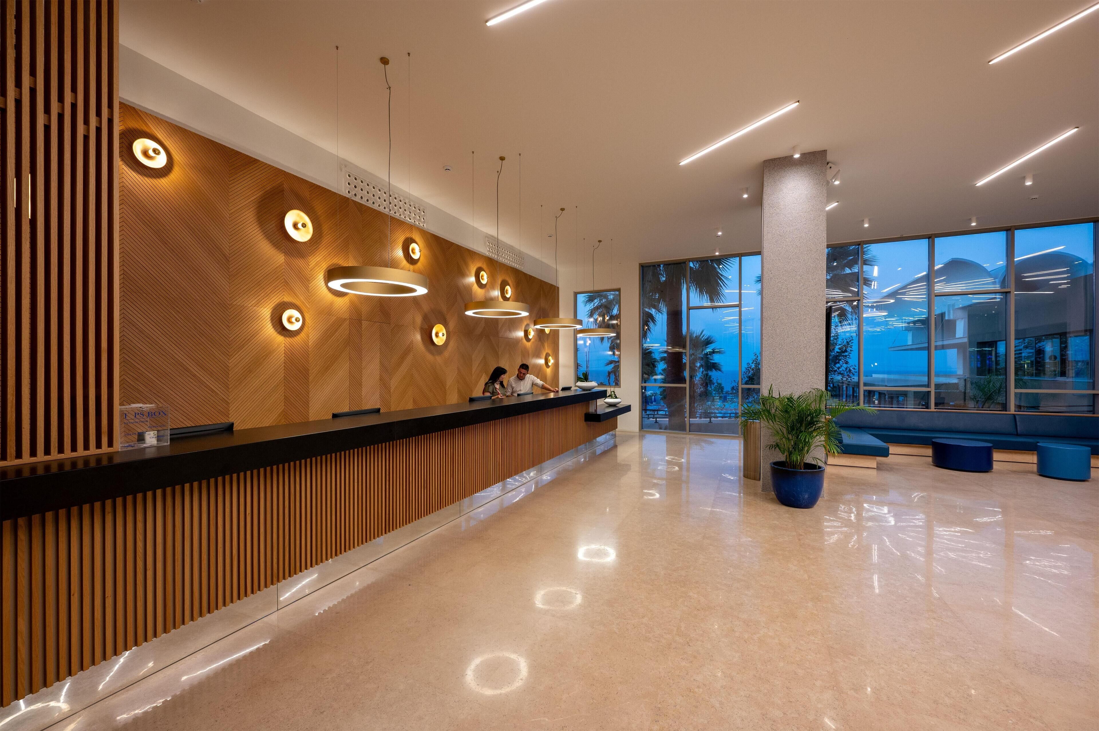 A modern reception area with a wooden panel wall, circular light fixtures, and a large window offering a view of palm trees.