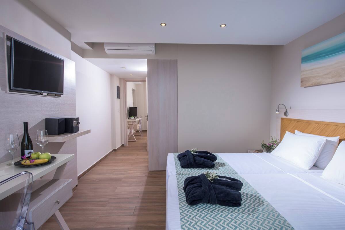 Una habitación de hotel moderna con una cama grande, un televisor de pantalla plana en la pared, un escritorio con computadora y una mesita con una botella y vasos.