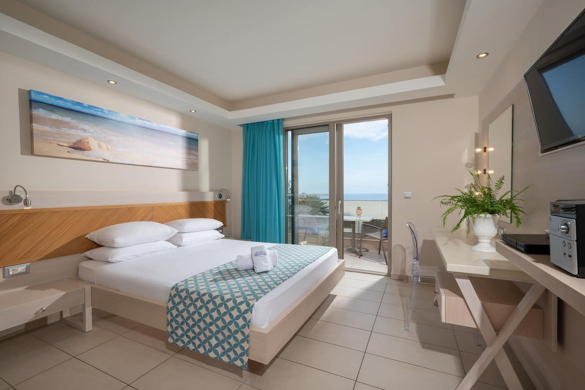 Una habitación de hotel con cama, escritorio, televisión y balcón con vista al mar.