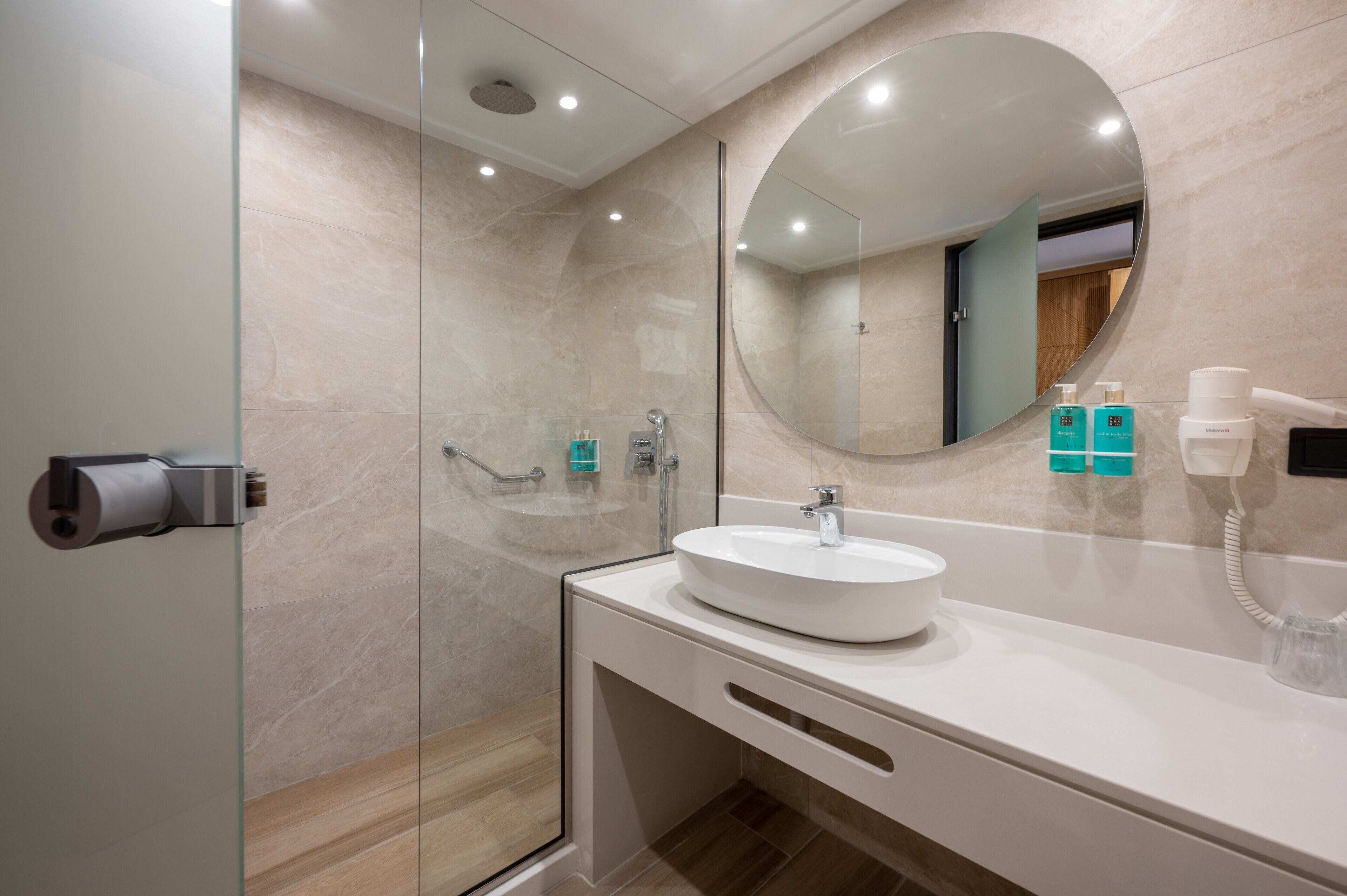 Een moderne badkamer met een douchecabine van glas, een ronde spiegel en een witte wastafel met ingebouwde telefoon.