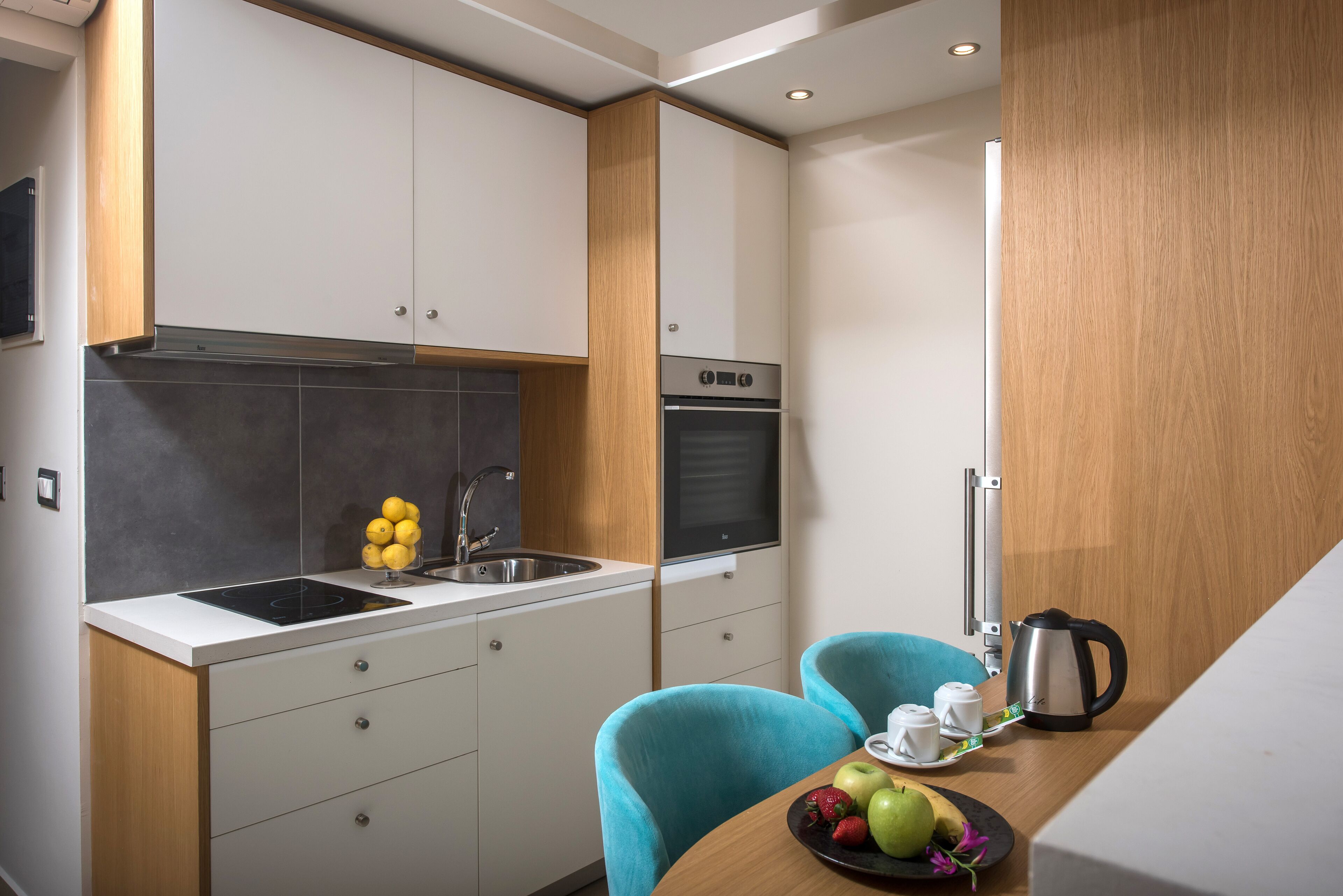A compact kitchen with a built-in oven, a sink, and a dining area with two chairs and a table set with fruits and a kettle.