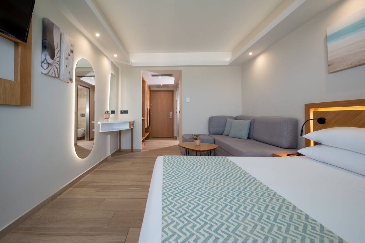 Una habitación de hotel moderna con una cama grande, un sofá, una mesita y un espejo, además se ve un baño a través de un arco.