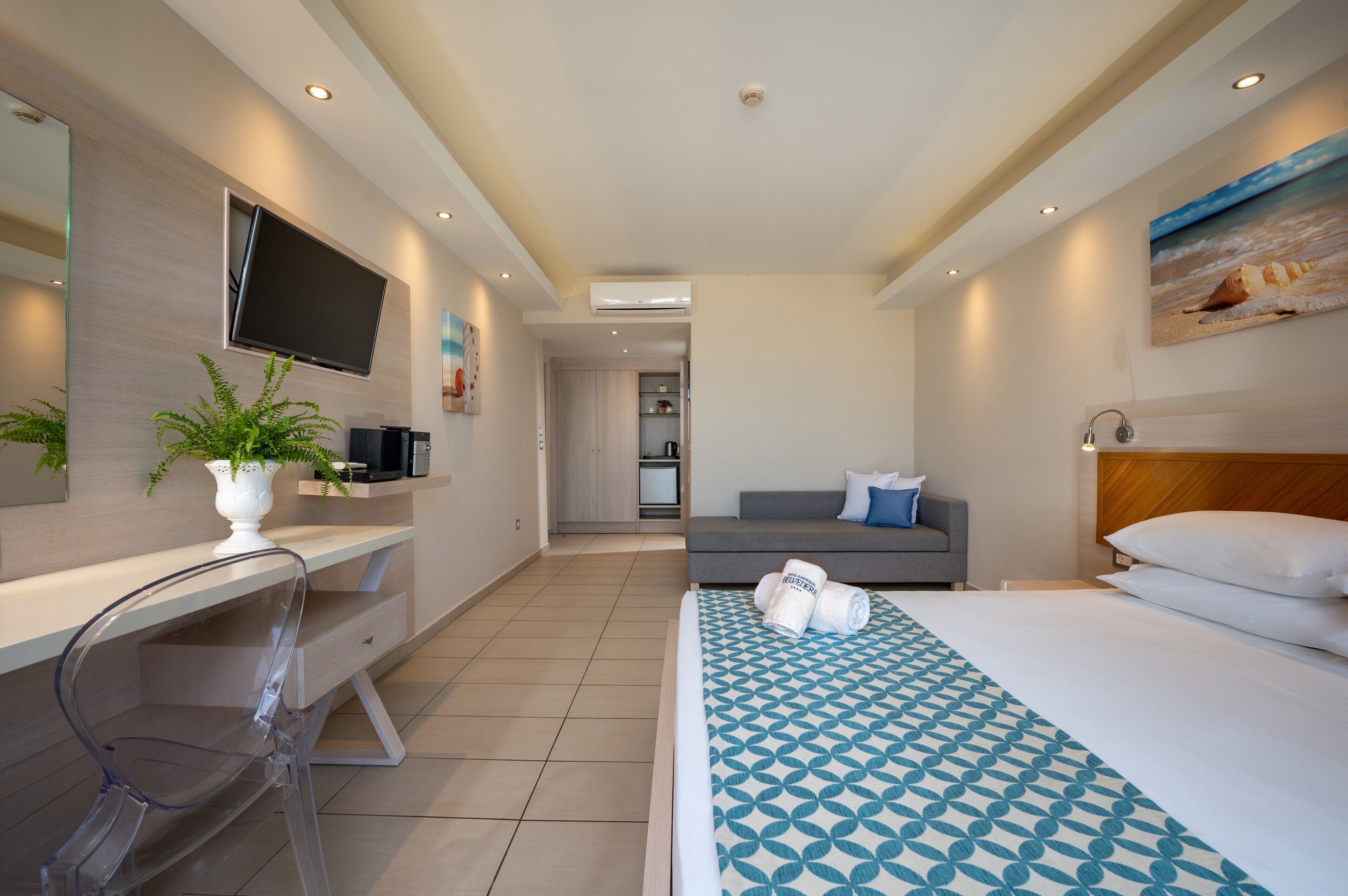 A modern hotel room with a flat-screen TV, a desk with a plant, a grey sofa, a bed with a patterned bedspread, and a painting on the wall.