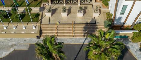 Vista aérea de un hotel con piscina central rodeada de palmeras, una escalera que conduce a la entrada y un edificio de diseño moderno con ventanas arqueadas y fachada estampada.