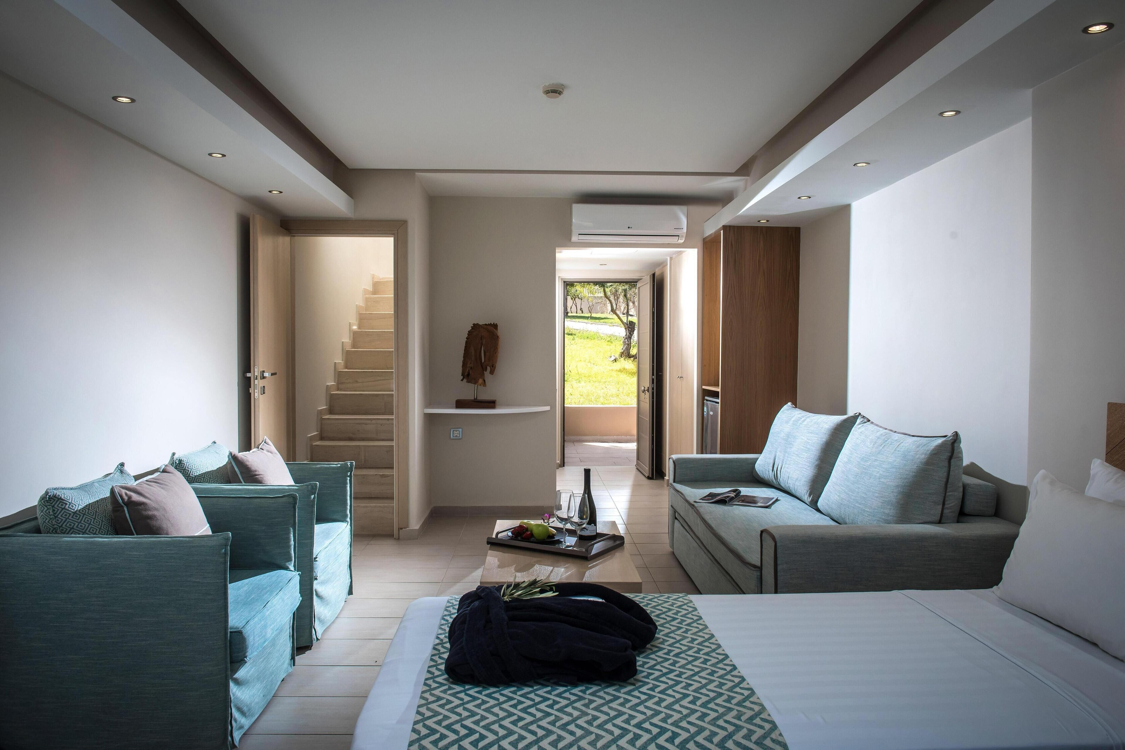 A modern hotel room with a bed, two sofas, a coffee table, and a staircase leading to a balcony.