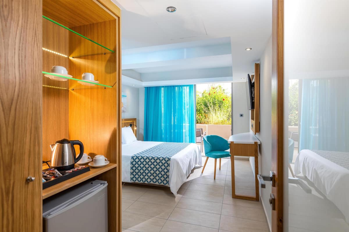 Una habitación de hotel con cama, escritorio, silla, kitchenette pequeña y balcón con cortinas azules.