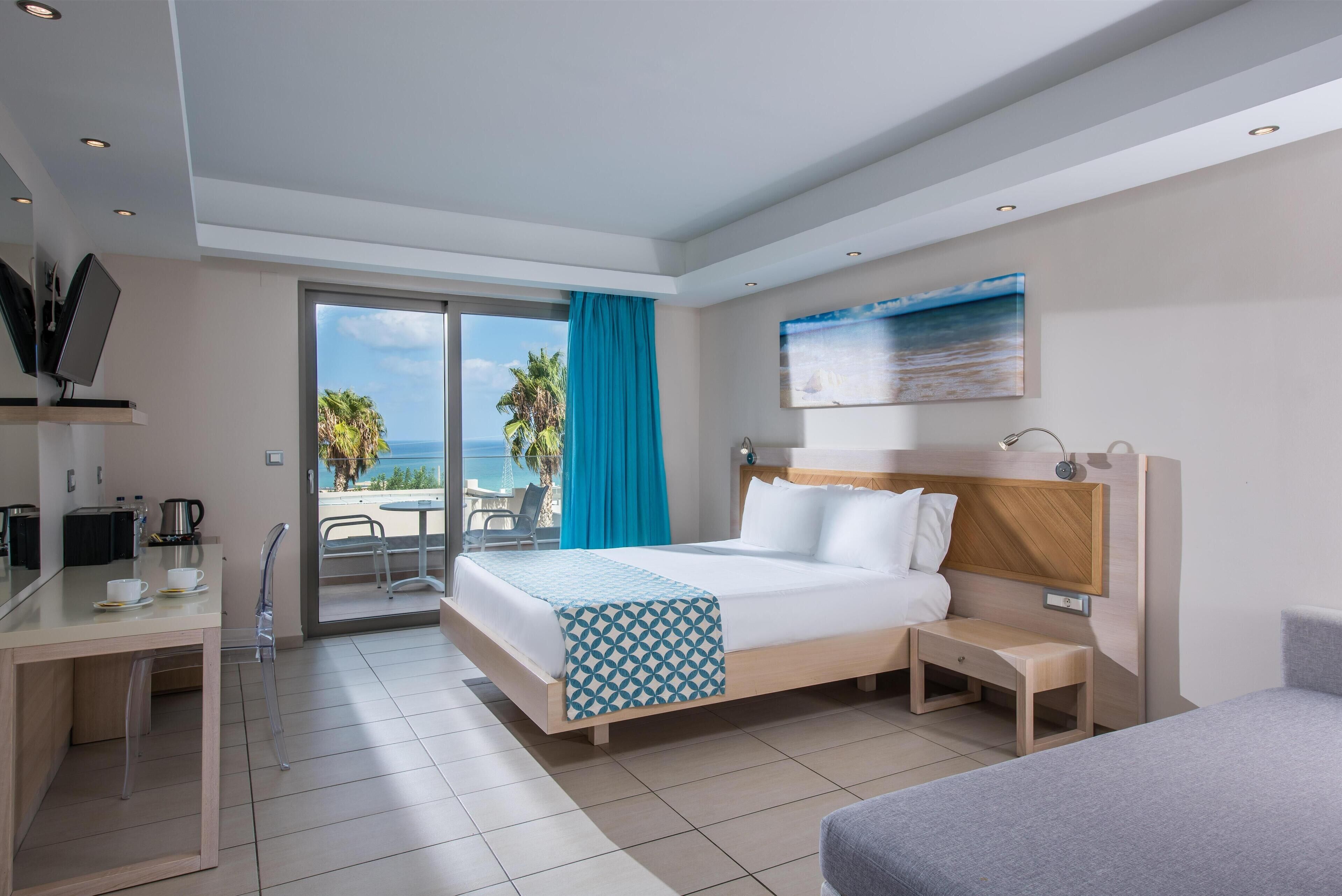 A modern hotel room with a bed, bedside table, and a desk with a chair. There is a large window with a view of the beach and palm trees.