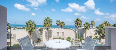 A balcony with a white table and two chairs, overlooking a beach with palm trees and a clear blue sky.