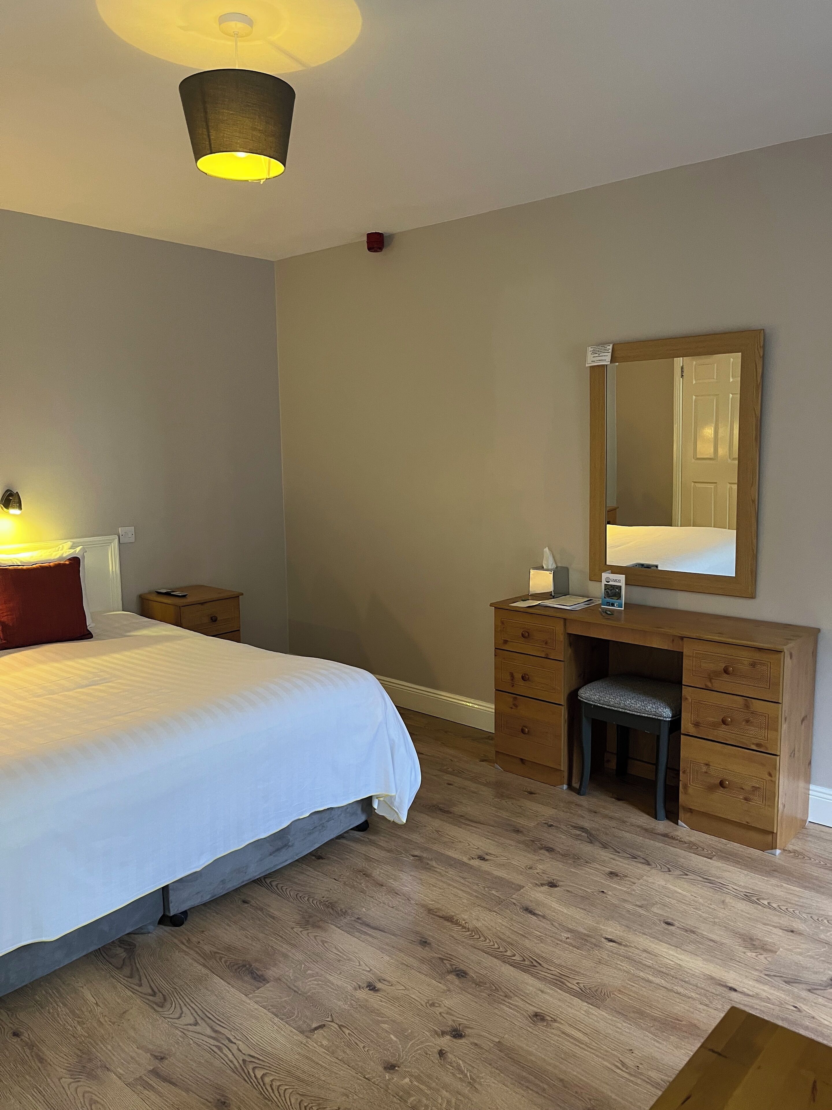 A bedroom with a bed, bedside table, lamp, dresser, mirror, and a wooden floor.