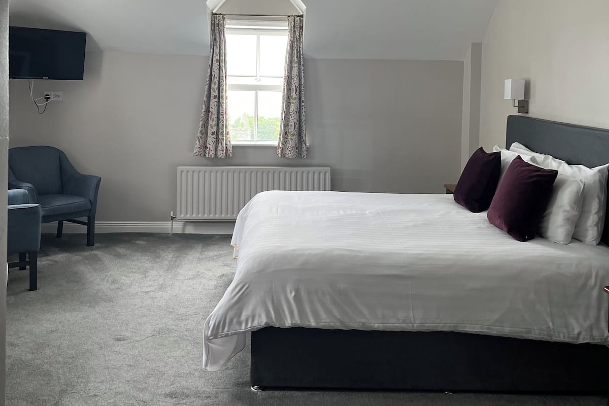 A bedroom with a bed, a chair, a TV, a window with curtains, and a radiator.