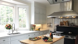 A modern kitchen with white cabinetry, a large island with fruit and wine, a stove, and a view of greenery through the windows. - Olallieberry Inn B&B (Cambria)