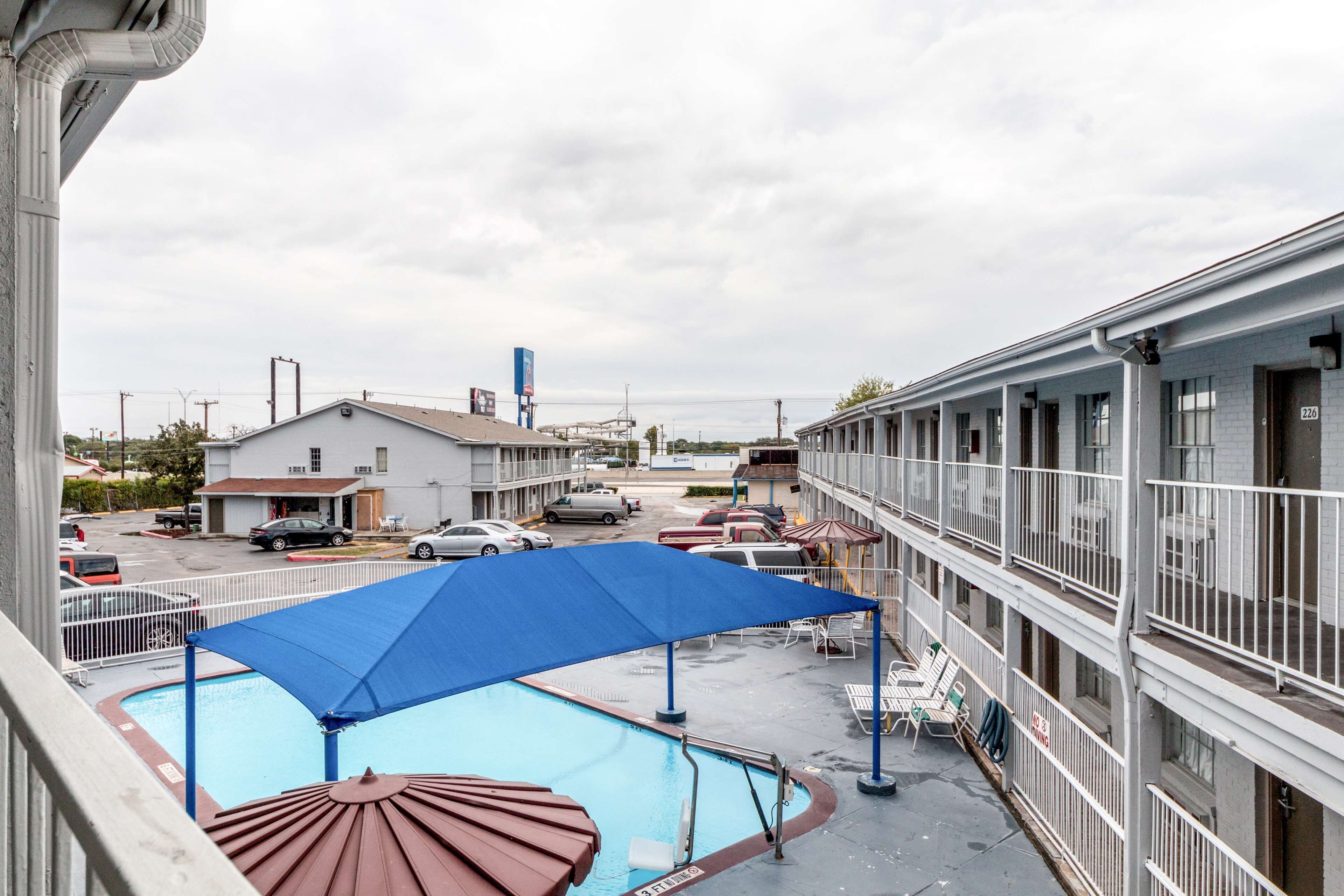 A hotel with a swimming pool, covered by a blue umbrella, and a parking lot with several cars.