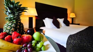 Una cama bien hecha con ropa blanca y un cabecero oscuro, un frutero con piña, fresas, uvas y una manzana, y dos lámparas de noche sobre una mesita.