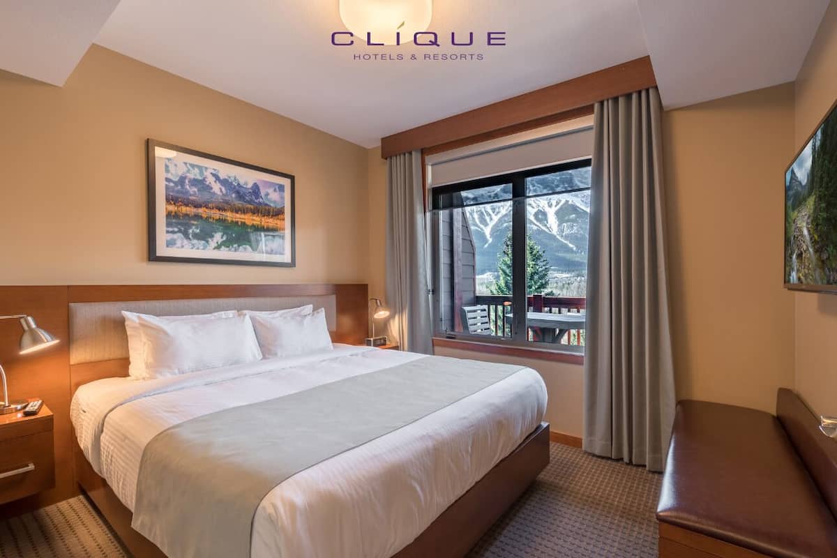 Una habitación de hotel con una cama grande, una mesita de noche, una lámpara de escritorio, una ventana con cortinas y un cuadro en la pared.