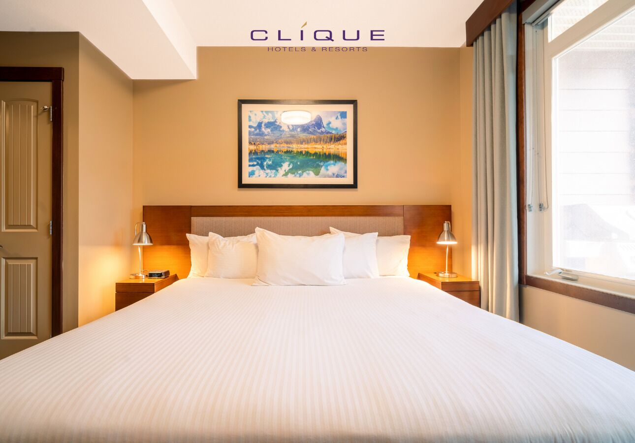 A hotel room with a large bed, two bedside lamps, a framed painting on the wall, and a window with curtains.