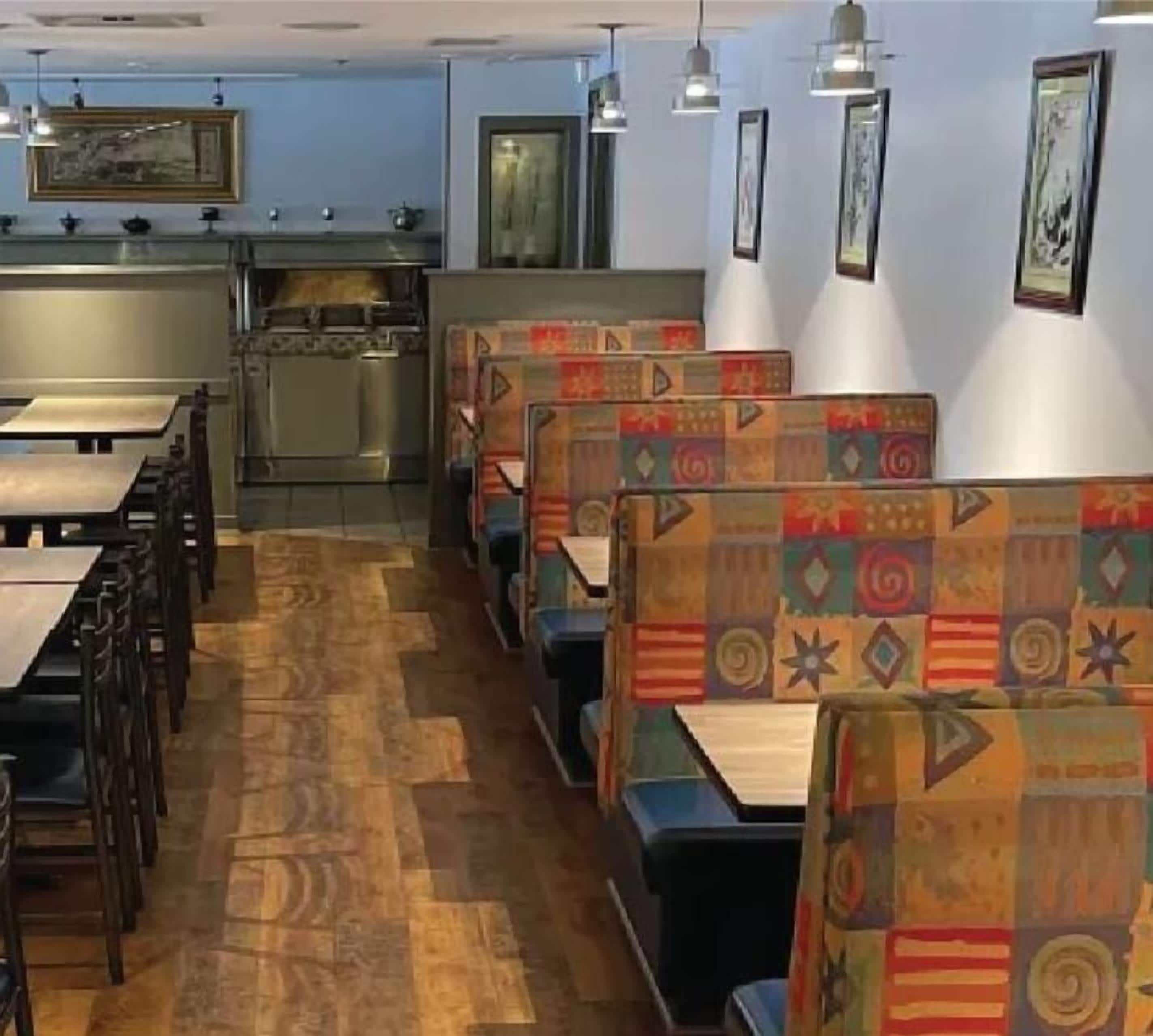 A restaurant interior with wooden flooring, booths with colorful upholstery, hanging pendant lights, and a kitchen area in the background.