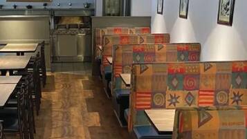 A restaurant interior with wooden flooring, booths with colorful upholstery, hanging pendant lights, and a kitchen area in the background.