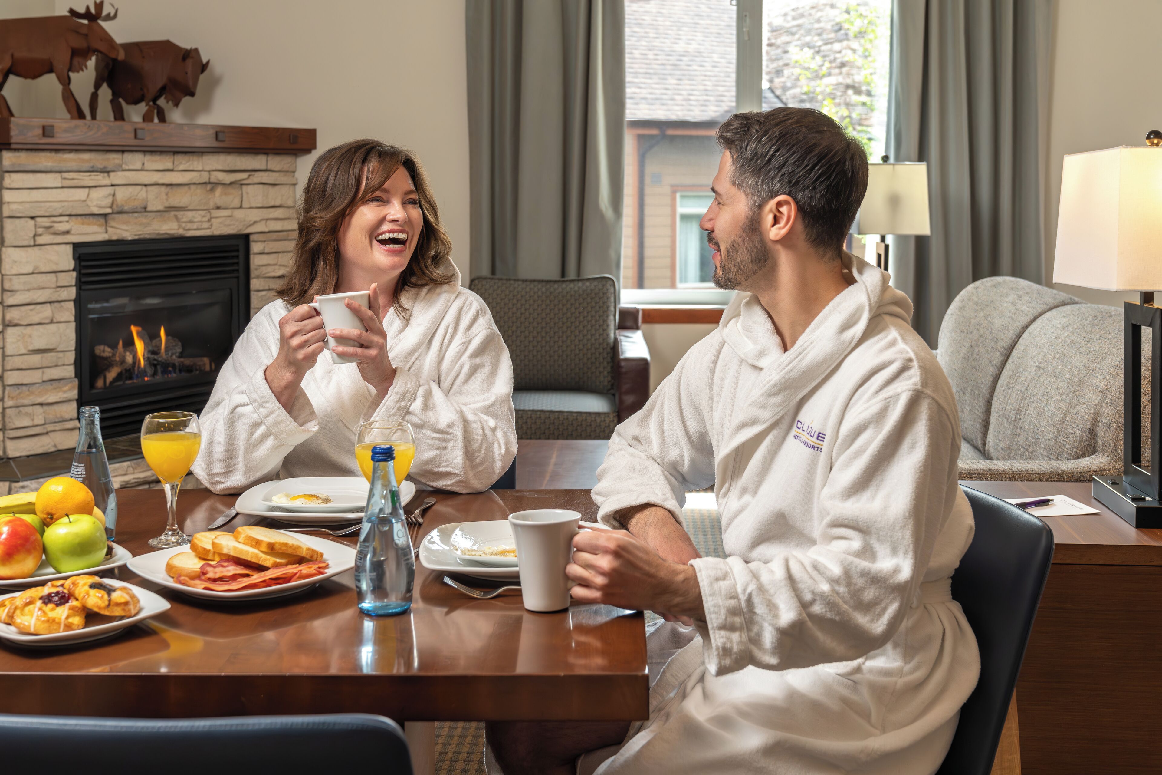 A couple sitting at a breakfast table with a fireplace, dining table, and a lamp in the background.