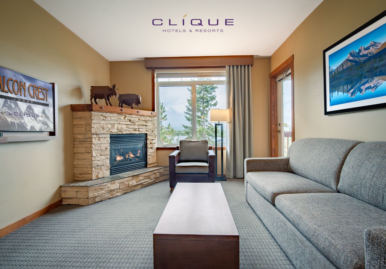 A hotel room with a stone fireplace, a sofa, a chair, and a large window offering a mountain view.