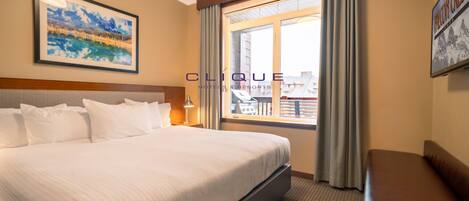 A hotel room with a large bed, a desk, a chair, a window with curtains, and a framed picture on the wall.