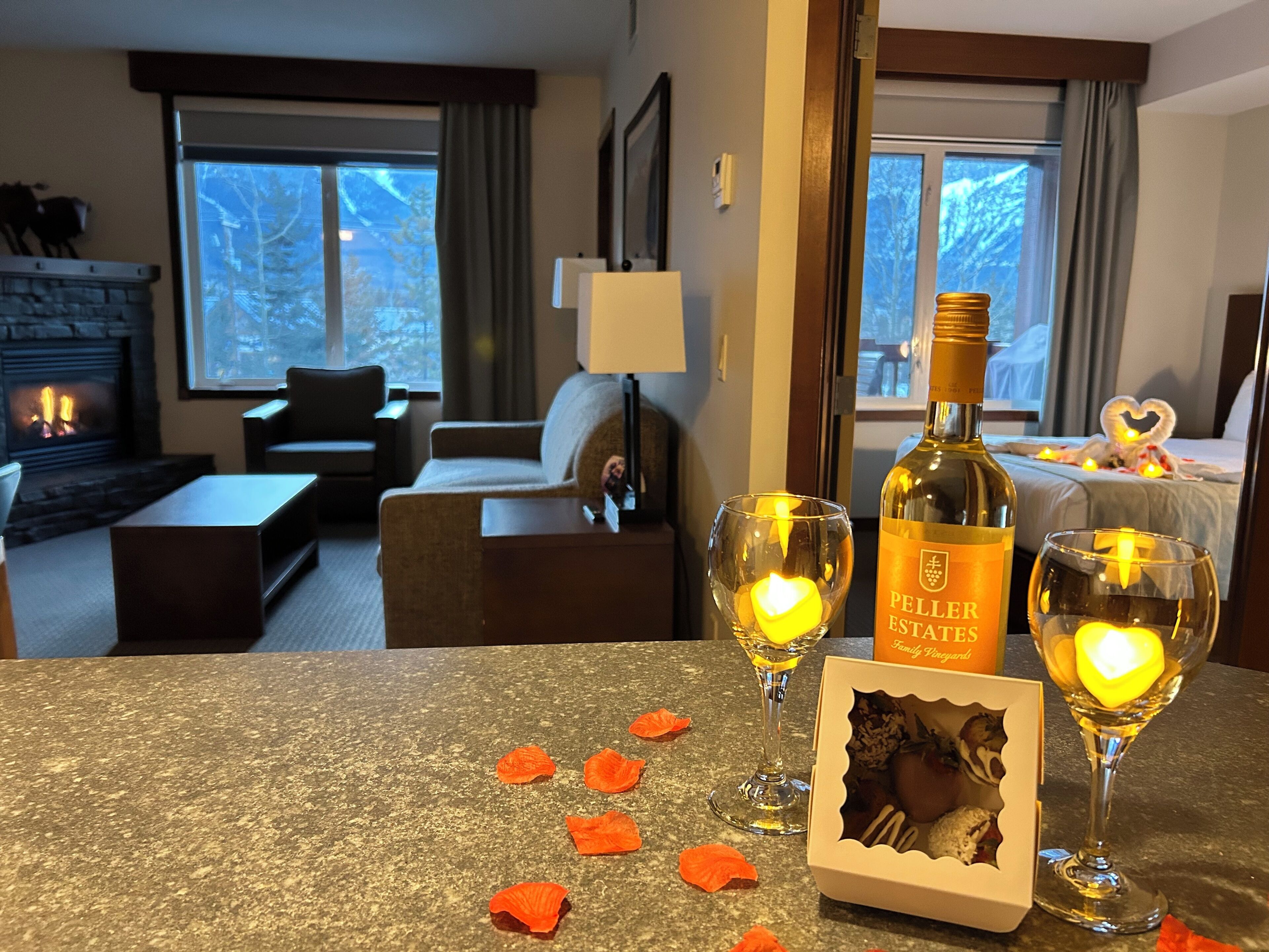 A hotel room with a fireplace, a sofa, a table with a bottle of wine, two glasses with candles, and a heart-shaped decoration on the bed.