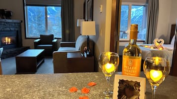 A hotel room with a fireplace, a sofa, a table with a bottle of wine, two glasses with candles, and a heart-shaped decoration on the bed.