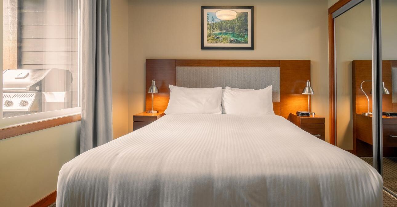 A hotel room with a large bed, two bedside tables with lamps, a window with curtains, and a framed picture on the wall.
