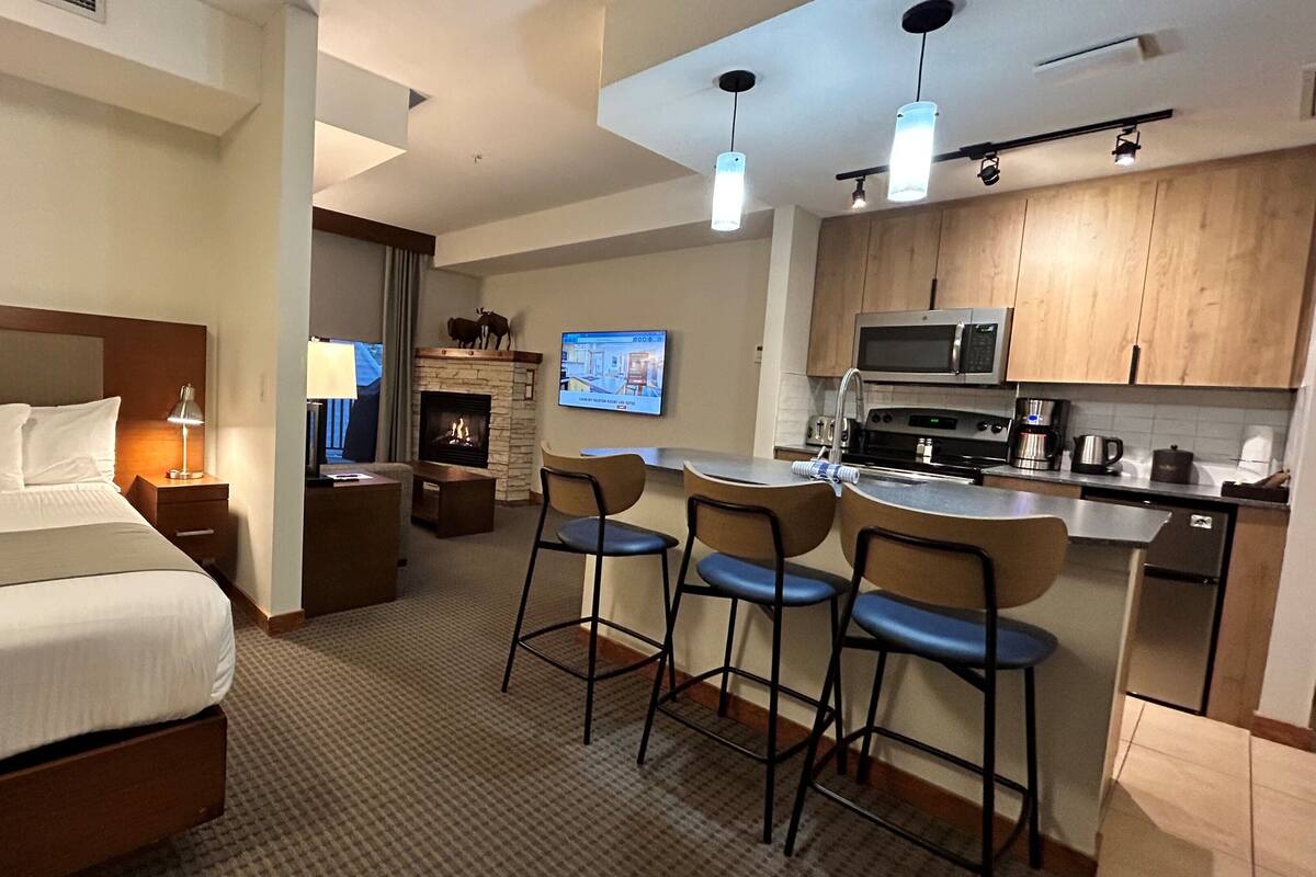 Habitación de hotel con cama, televisión, chimenea, espacio de cocina con microondas y barra con taburetes.