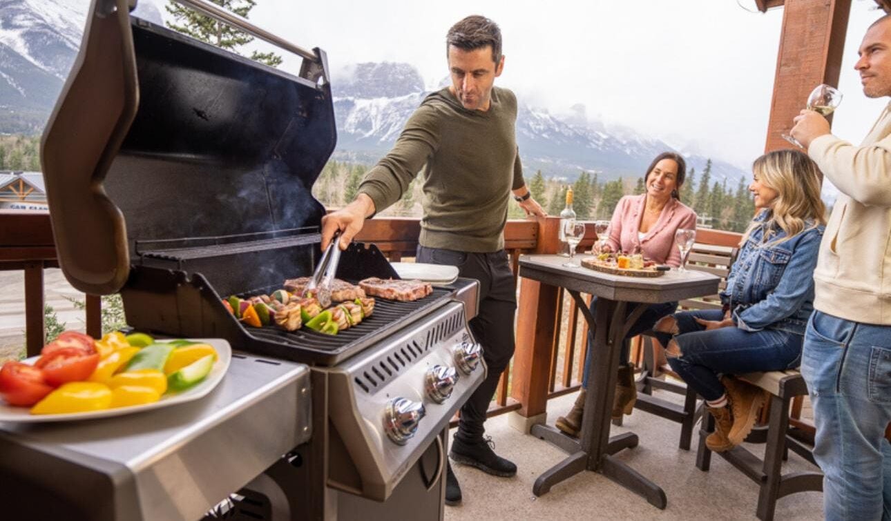 A barbecue grill with vegetables and meat, a table with drinks, and a mountainous backdrop.