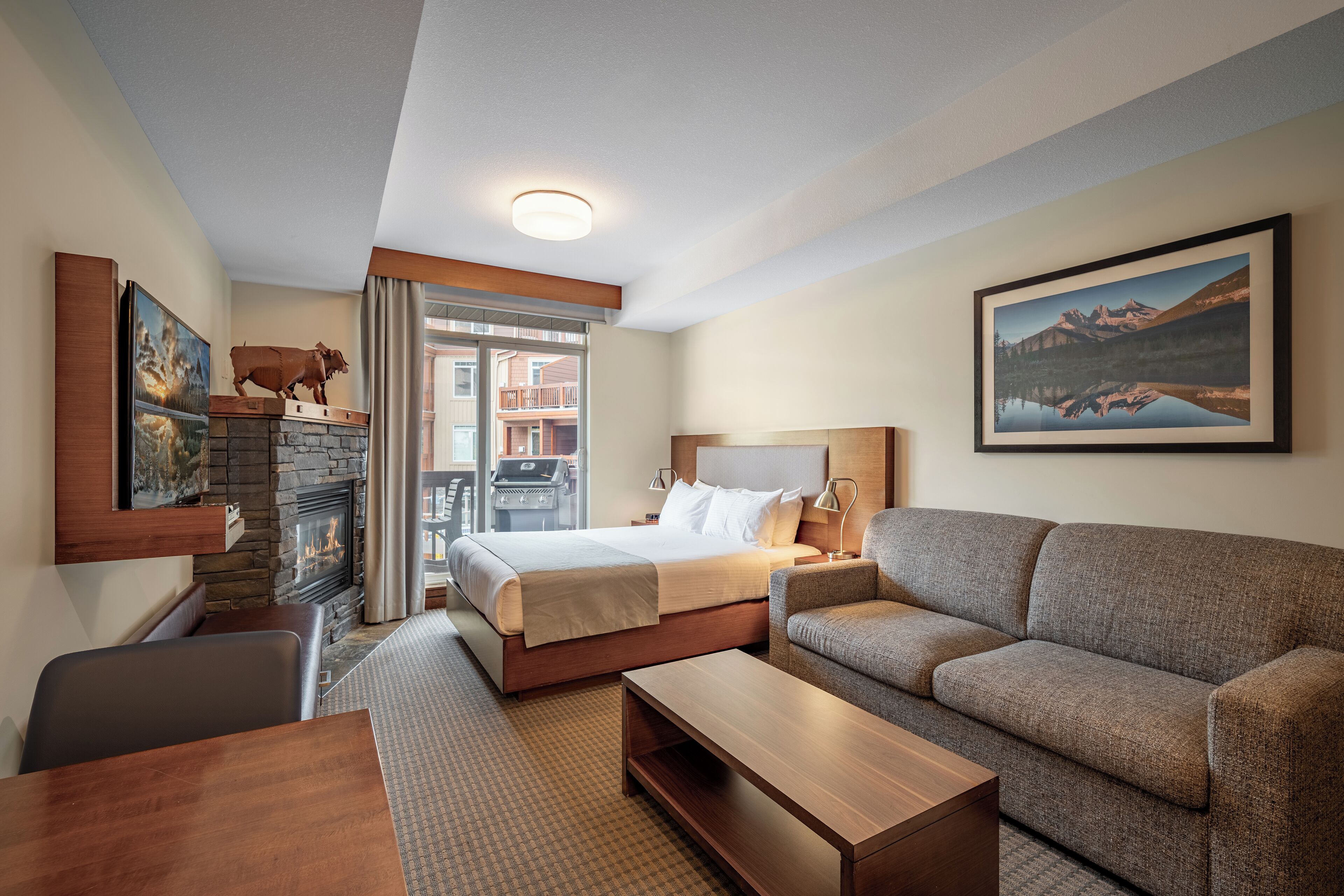 A hotel room with a bed, a sofa, a desk, a fireplace, and a balcony with a view of a mountain.