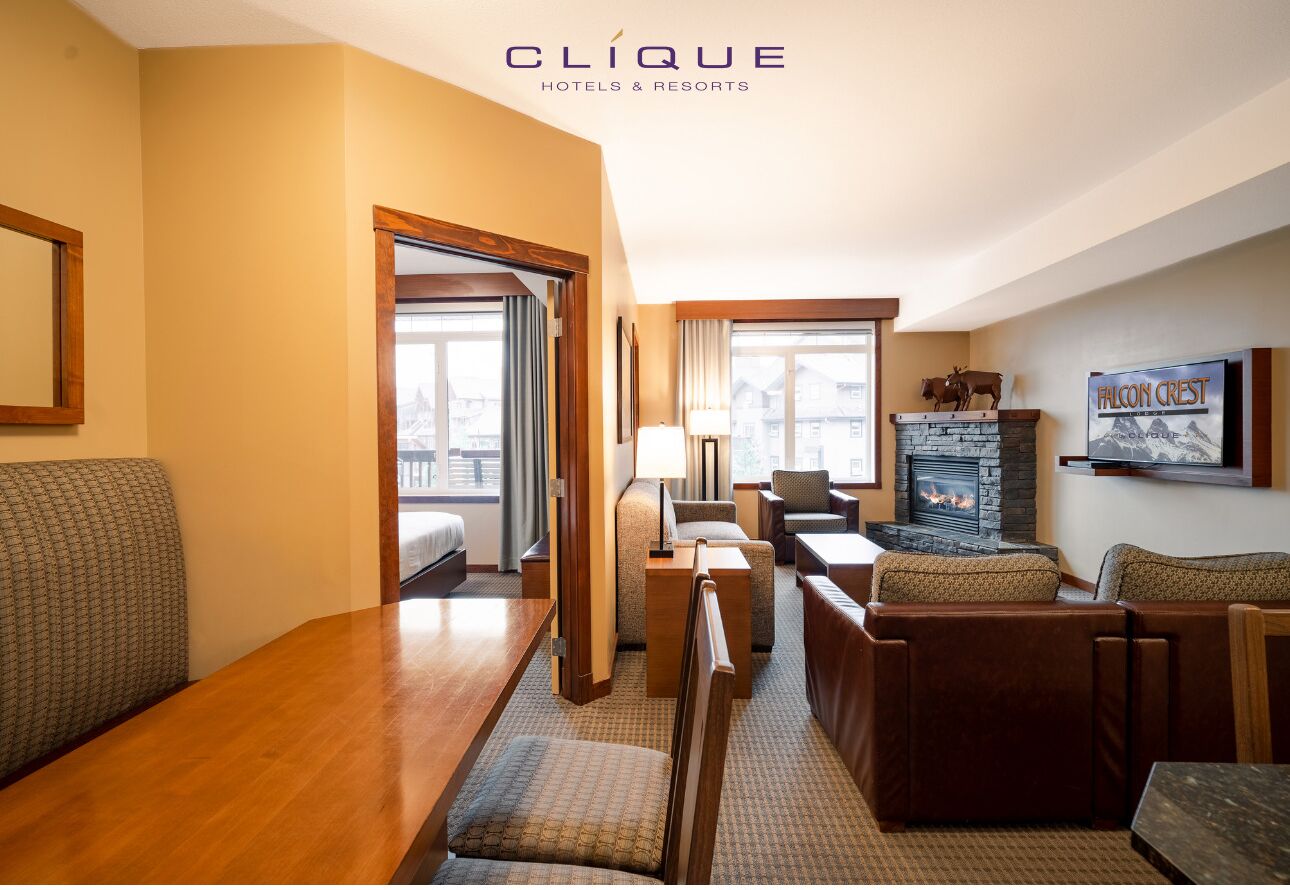 A hotel room with a wooden floor, a sofa, a dining table, a fireplace, and a view of the city through large windows.