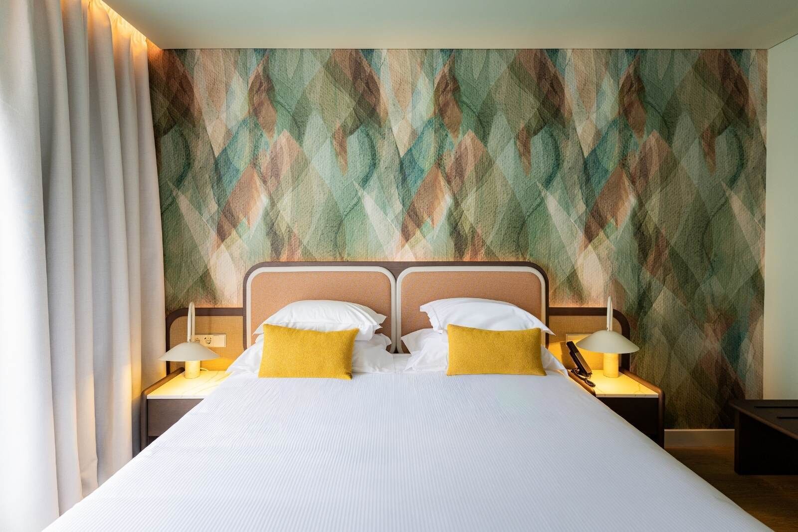 A double bed with white bedding and two yellow pillows, flanked by nightstands with lamps, in a room with a geometric patterned wall.