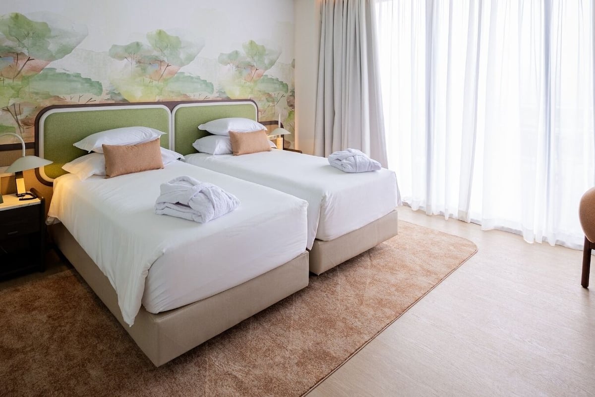Una habitación de hotel con dos camas, una mesita de noche, una lámpara, una silla y una ventana con cortinas.