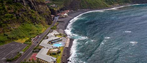 A coastal hotel with a swimming pool, a parking lot, and a road leading to the beach.