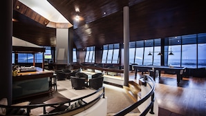 Amplia sala de estar con disposición curvilínea de asientos, ventanas grandes con vistas al océano y un alto techo con paneles de madera.