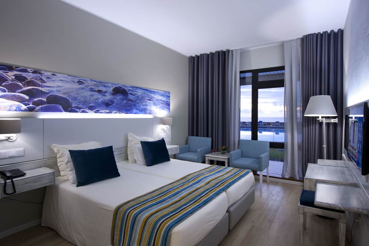 A modern hotel room with a large bed, a desk, a chair, and a view of the city.