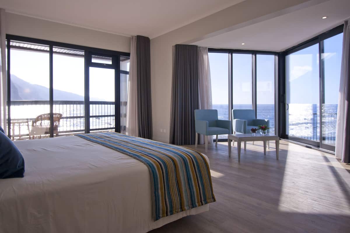A hotel room with a large bed, a balcony with a view of the sea, and a seating area with two chairs.