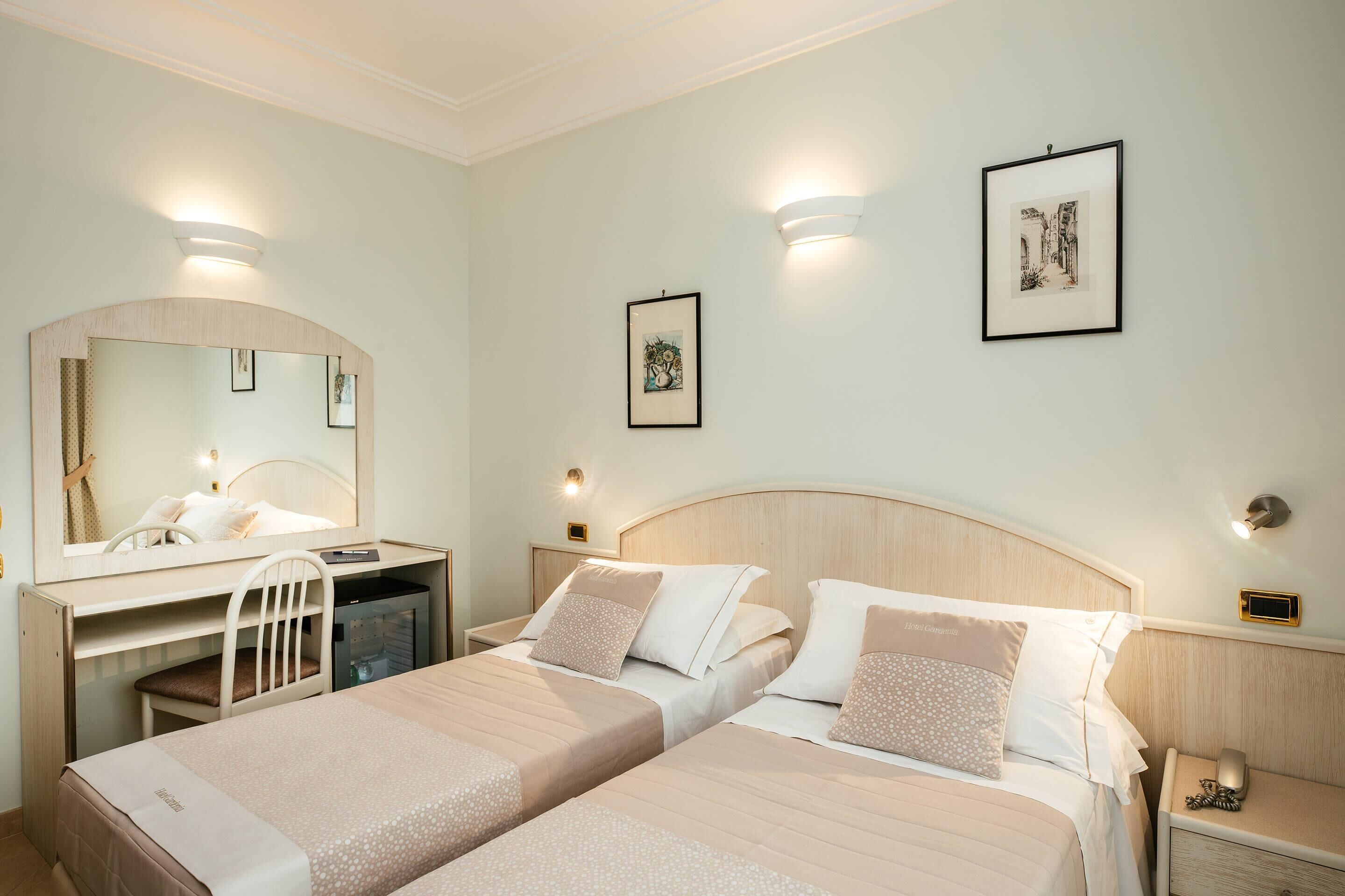Ein Hotelzimmer mit zwei Betten, einem Schreibtisch, einem Stuhl, einem Spiegel und Bildern an den Wänden.