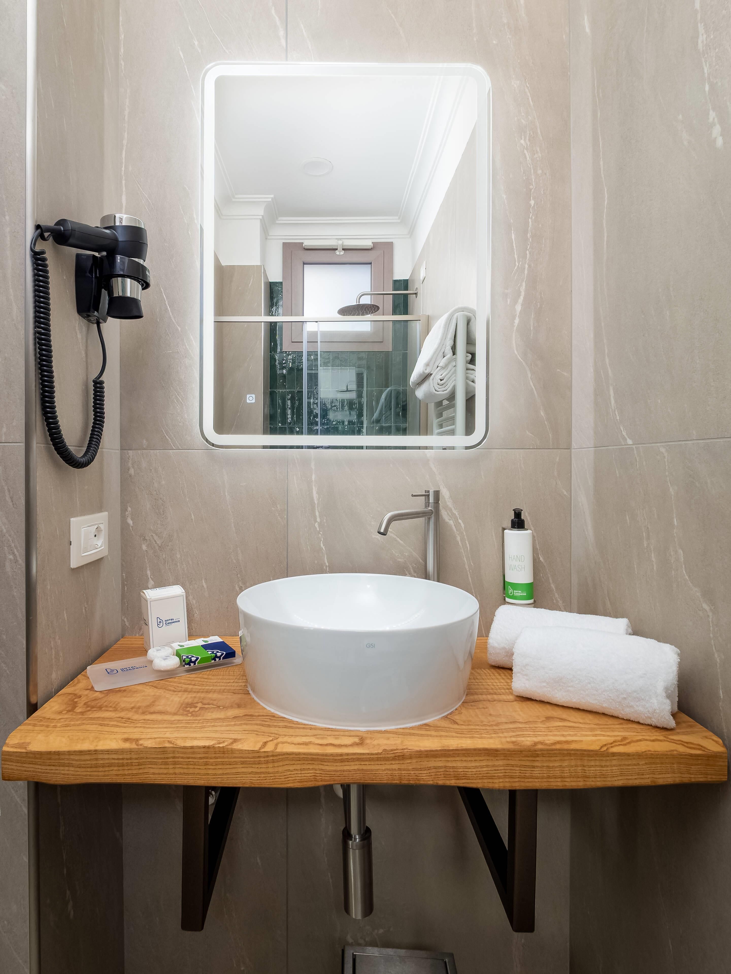 A bathroom with a wooden vanity, a white sink, a handheld showerhead, a hairdryer, and a hand wash bottle.