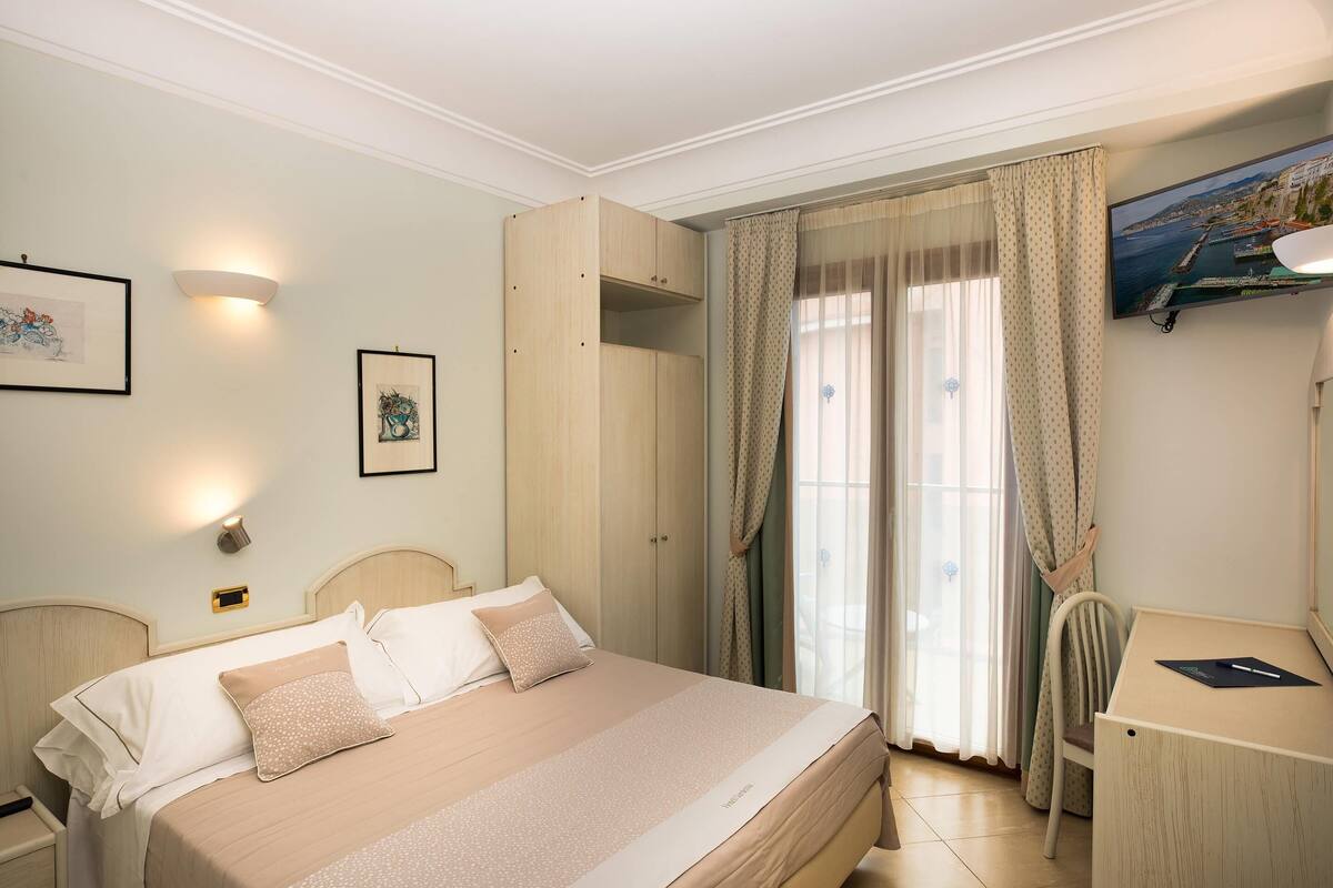 Une chambre bien rangée, avec un lit, un bureau et une chaise. Il y a des rideaux à la fenêtre et des œuvres d’art accrochées aux murs.