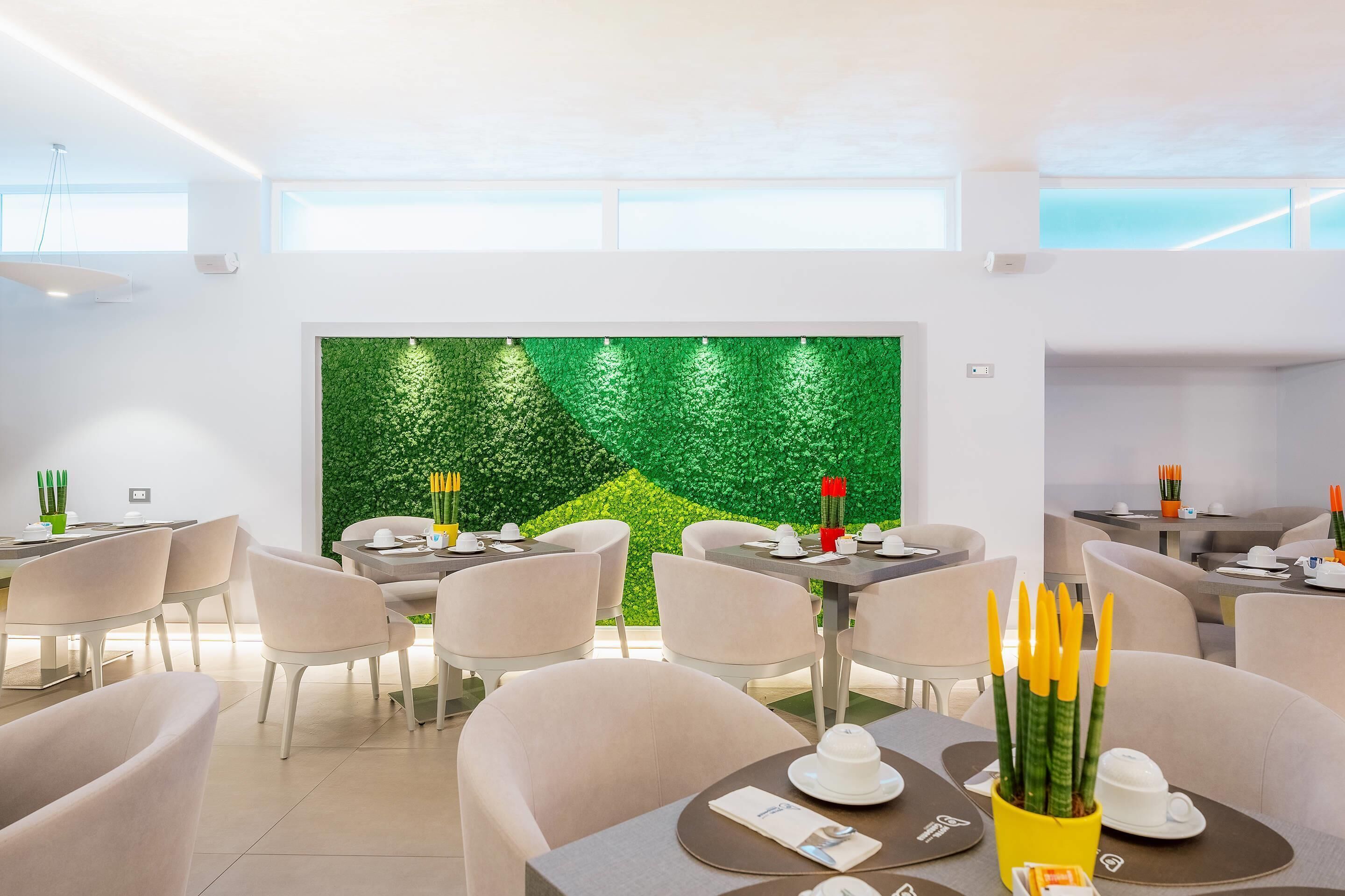 A modern dining area with tables and chairs, a green wall with a leafy pattern, and a bright ceiling.