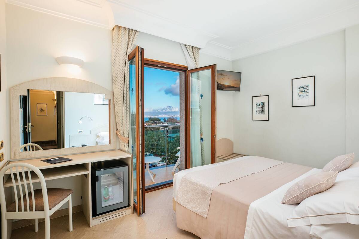 Une chambre d’hôtel comprenant un lit, un bureau, une chaise, un petit réfrigérateur, une fenêtre donnant sur l’extérieur et une porte menant à un balcon.
